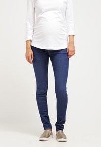 Kvinna som bär en vit långärmad skjorta, mörkblå tajta jeans och metallic slip-on skor, som står mot en enkel vit bakgrund.