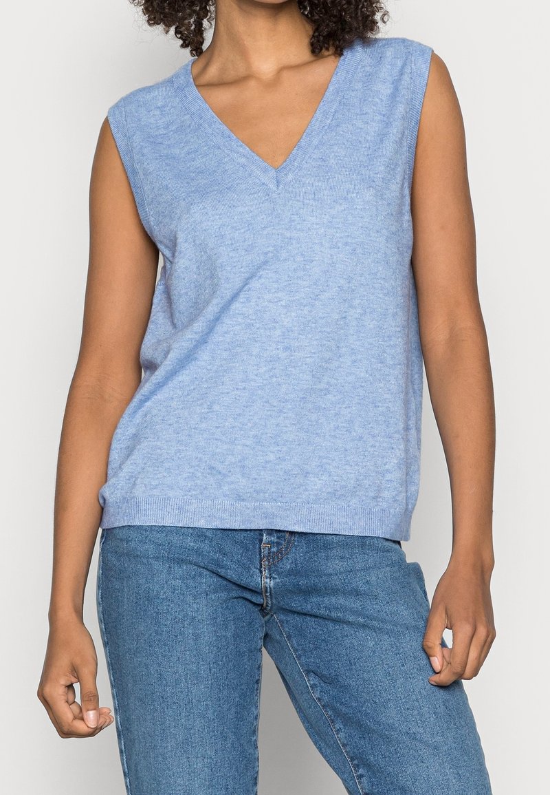 Femme portant un gilet en tricot bleu clair sans manches à col en V et un jean bleu en denim, debout devant un fond uni.