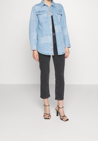 Světle modrá džínová bunda s dvěma náprsními kapsami a knoflíkovým zapínáním, nošená přes černý top, doplněná černými zkrácenými kalhotami a sandály na vysokém podpatku.