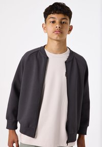 Chaqueta de cremallera gris oscuro con puños acanalados, textura suave y cuello estándar, superpuesta a una camiseta de color beige claro.