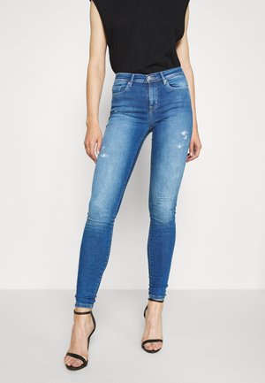 Woman wearing high-waisted blue skinny jeans with slight distressing and black strappy heels, paired with a black sleeveless top.