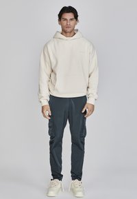 Cremefarbener Hoodie mit einer Fronttasche, kombiniert mit dunkelblauen Cargo-Hosen mit Seitentaschen und weißen Sneakers. Einfarbiger Hintergrund.