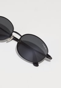 Black sunglasses with oval lenses, featuring a glossy frame and silver accents. The brand "vogue" is printed in white on the frame.