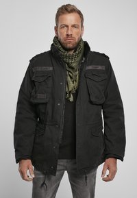 Zwarte militaire stijljack met meerdere zakken aan de voorkant, ritssluiting en verstelbare trekkoorden. Ggedragen over een donkere gebreide trui, geaccessorieerd met een groene sjaal.