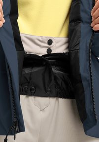 Marine Leichtjacke, geöffnet, zeigt schwarzen Futter und elastischen Bund mit Druckknöpfen; getragen über einem gelben Fleece-Oberteil und beigen Hosen.