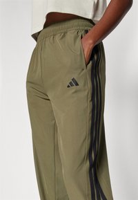 Olivengrønne træningsbukser med elastisk talje, der har to sidelommer og sorte striber ned langs siderne. Adidas-logo i sort.