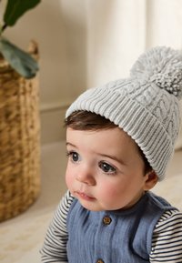 Lysegrå strikket hue med en pompon, som har et kabelstrikket mønster. Barnet bærer et blåt tøj med knapper og stribede ærmer.