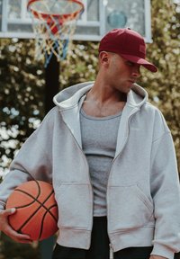 Gray zip-up hoodie over a gray tank top, black shorts, holding an orange basketball; wearing a maroon cap with perforated design.