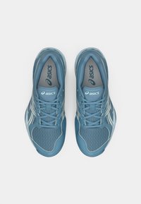 Blå nettstoff treningssko med grå detaljer, med avrundet tå, polstret tunge og flate lisser. Merke synlig på tungen.