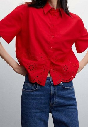 Kobieta ubrana w jasnoczerwoną koszulę na krótki rękaw z haftowanym wykończeniem z ażurowym wzorem, zestawioną z wysokim stanem niebieskich dżinsów.