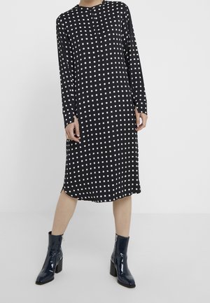 Schwarzes Midikleid mit weißem quadratischem Muster, langen Ärmeln und rundem Ausschnitt, getragen mit glänzenden schwarzen Stiefeletten mit Blockabsätzen.