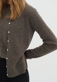 Moteris dėvi rudą vilnonį kardiganą su sagomis ir ribotu apačiu bei juodas kelnes, stovinti su rankomis ant klubų priešais paprastą foną.