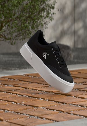 Fekete sportcipő fehér "CK" logóval az oldalon, vastag fehér talppal és fekete fűzőkkel, egy kültéri fa csempe és kavicsos padló fölött elhelyezve.