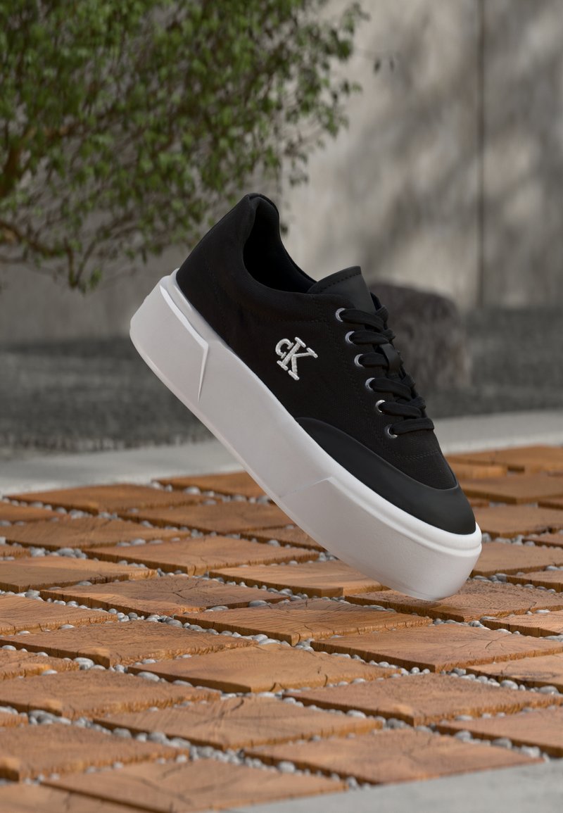 Μαύρο αθλητικό παπούτσι με λευκό λογότυπο "CK" στη πλευρά, χοντρή λευκή σόλα και μαύρα κορδόνια, τοποθετημένο πάνω σε ξύλινο πλακάκι και δάπεδο με βότσαλα σε εξωτερικό χώρο.