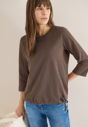 Kvinde iført en brun langærmet top med knyttet kant og blå jeans, der holder en hvid jakke mod en ensfarvet lys væg som baggrund.