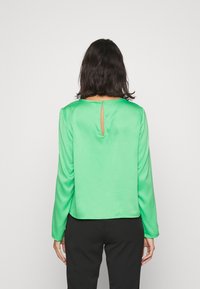 Top lungo a maniche con scollo rotondo in satin verde, con un piccolo oblò sul retro. Tessuto liscio e vestibilità rilassata, abbinato a pantaloni neri.