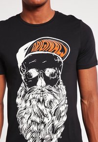 Camiseta negra con un diseño gráfico blanco de una calavera con gafas de sol y una larga barba, acentuada por una gorra naranja y blanca.