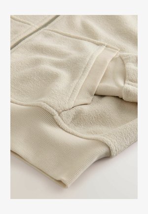 Hanorac din fleece bej cu manșete și tiv ribbate, având o textură moale și un fermoar neted în contrast. Straturile de țesătură creează dimensiune.