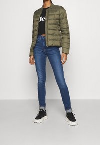 Persona vestida con una chaqueta acolchada verde oliva, una camiseta negra corta, jeans ajustados azules, calcetines grises y zapatillas chunky en blanco y negro.