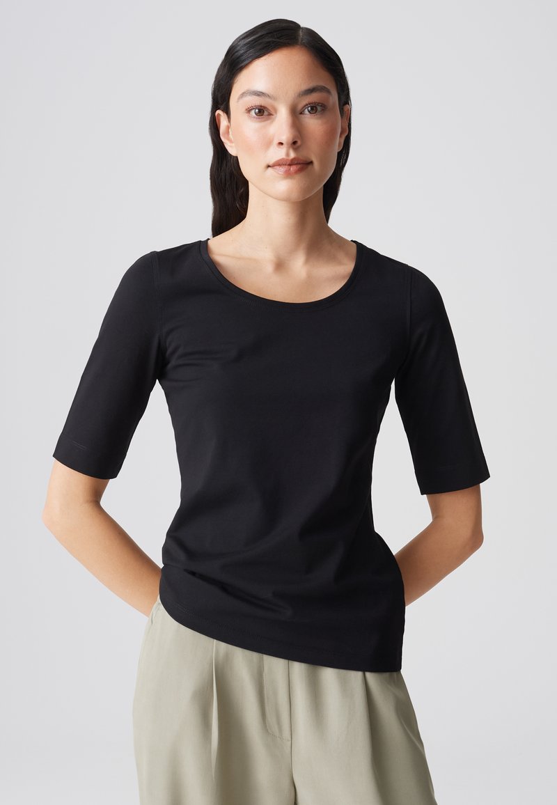 Femme aux longs cheveux foncés portant un t-shirt noir moulant à manches courtes et un pantalon beige, debout les mains dans le dos.