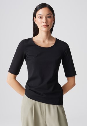 Vrouw met lang donker haar, gekleed in een nauwsluitend zwart T-shirt met korte mouwen en beige broek, staand met haar handen achter haar rug.