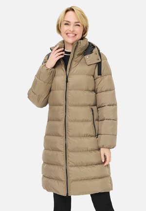 Frau lächelt und trägt einen beigefarbenen, knielangen Steppmantel mit Reißverschluss, Taschen und abnehmbarer Kapuze über schwarzer Hose vor weißem Hintergrund.