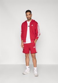 Raudonas sportinis aprangos komplektas, sudarytas iš užtrauktuku užsegamo džemperio, balto marškinėlio ir šortų su baltomis juostelėmis ir logotipu. Avima balti sportbačiai.