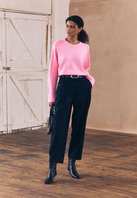 Suéter de punto rosa brillante con mangas largas, combinado con pantalones plisados azul marino y botines negros de tacón, sosteniendo un pequeño bolso de mano negro.