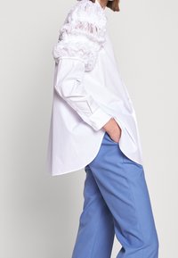 Witte blouse met geplooide, gerimpelde mouwen; gecombineerd met lichtblauwe broeken. De blouse heeft een afgeronde zoom en knoopdetails op de manchetten.