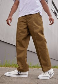 Khaki kalhoty s širokými nohavicemi vyrobené z odolného materiálu, kombinované s bílými teniskami s černým hvězdným vzorem, prezentovány na dlážděném povrchu.