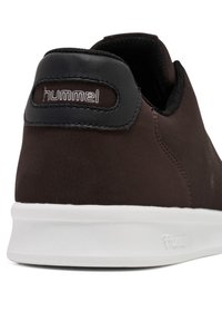 Brązowy sneaker o gładkiej teksturze i białej gumowej podeszwie. Posiada czarny akcent na pięcie z białym logo "hummel".