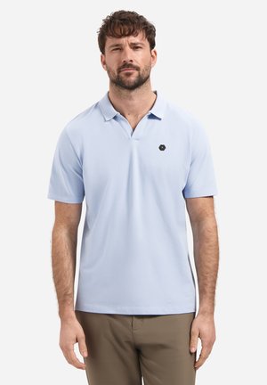 Man met kort bruin haar, gekleed in een lichtblauw poloshirt met korte mouwen en kraag, en een bruine broek, staand tegen een witte achtergrond.