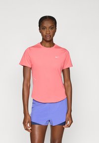 Γυναίκα που φοράει αθλητική μπλούζα Nike σε κοραλλί ροζ με κοντά μανίκια και μπλε αθλητικό σορτς, στέκεται μπροστά σε απλό φωτεινό φόντο.