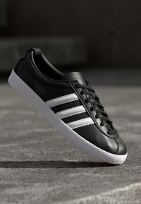 Sapatilhas de couro preto com três riscas brancas, atacadores planos pretos e uma sola de borracha branca texturizada. Design clássico de cano baixo.