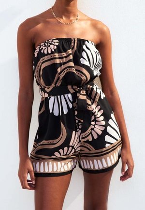 Femme portant une combinaison noire bustier avec des motifs floraux et abstraits beige et blanc, nouée à la taille avec une ceinture en tissu assortie.