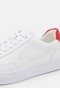 Sneaker bianca in pelle con accentuazione rossa sul tallone, lacci bianchi piatti, logo in rilievo sul lato e suola in gomma testurizzata.