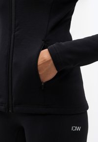 Χέρι στην πλαϊνή τσέπη με φερμουάρ ενός μαύρου μπουφάν με φερμουάρ, φορεμένο πάνω από μαύρο κολάν με λευκό λογότυπο ICIW στο ισχίο.