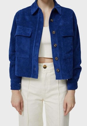Woman wearing a cropped blue corduroy button-up jacket over a white crop top and high-waisted off-white pants with a brown button.