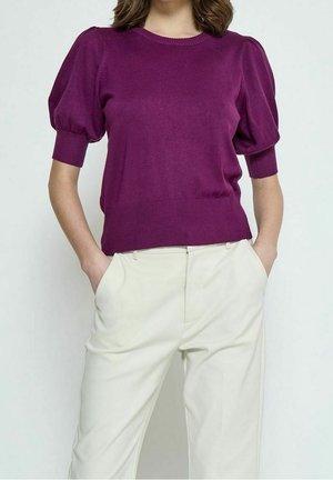 Femme portant un haut bouffant violet à manches courtes et un pantalon couleur crème, les mains dans les poches, sur un fond clair.
