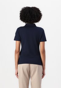 Marineblå polo skjorte med korte ermer og krage, laget av glatt stoff, sett fra baksiden, kombinert med lyse beige bukser.