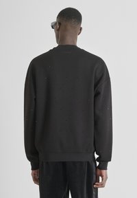 Felpa nera con superficie testurizzata, caratterizzata da piccoli accenti metallici. Vestibilità oversize con polsini e orlo a coste. Nessun logo visibile sul retro.