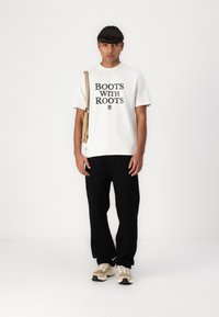 Fehér pamut póló "BOOTS WITH ROOTS" felirattal, laza fekete nadrággal és világos részletekkel, valamint hálós betétekkel ellátott bézs sneakerrel párosítva.