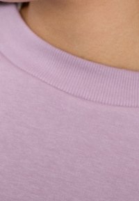 Sudadera de lavanda clara con escote acanalado. La tela parece suave y ligeramente texturizada, mostrando un sutil patrón jaspeado.