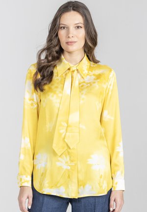 Kobieta o długich falowanych włosach, ubrana w żółtą jedwabną bluzkę w kwiaty z wiązaniem na szyi, w połączeniu z niebieskimi dżinsami, stojąca na jasnym tle.