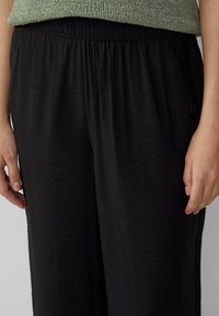 Mains reposant à côté d'un pantalon noir texturé lâche avec une taille élastique, associé à un haut en tricot vert.