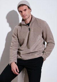 Bež flis pulover s polu-zip ovratnikom, dugim rukavima i teksturiranim materijalom. Nošen s crnim hlačama i sivim pletenim beanie šeširom.