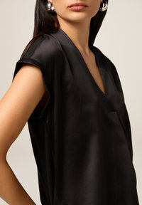 Top in satin nero con scollatura a V, maniche corte e vestibilità ampia, caratterizzato da una leggera drappeggiatura per un effetto texture e un aspetto morbido.