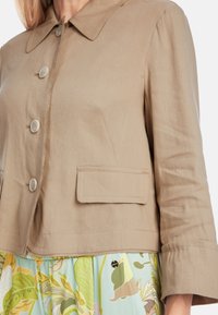 Light beige cropped jacket made of textured fabric, featuring a pointed collar, four buttons, and a single front pocket.