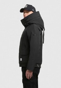 Juodas izoliuotas švarkas su aukštu apykakle, priekiniais užtrauktukais, didelėmis šoninėmis kišenėmis ir raibais rankogaliais. Dėvimas su juodu kepurėle.