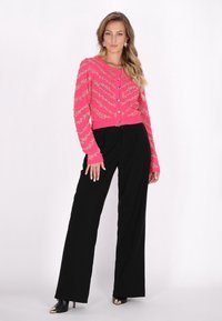 Cardigan rosa lavorato a maglia con decorazioni dorate, maniche lunghe e chiusura a bottoni, abbinato a pantaloni larghi neri e stivali alla caviglia.